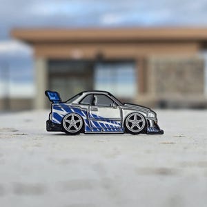 May include: A silver enamel pin shaped like a sports car featuring a blue and white graphic stripe pattern along the side, a rear spoiler, and five-spoke wheels. The car has a low-profile design with a front air dam and detailed window outlines.