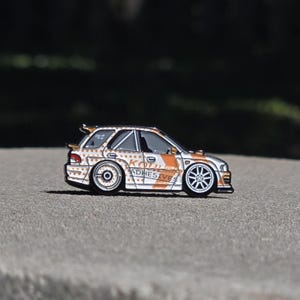 May include: A metal enamel pin shaped like a white rally car with orange polka dots and orange diagonal stripes. The side of the car features the text KOHLER ADHESIVES in orange lettering. The car has a rear spoiler and silver multi-spoke wheels.