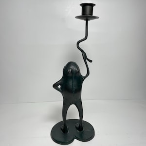 May include: A dark green metal candlestick holder in the shape of a frog. The frog figure is holding up a curved stem with a round candle holder at the top. The base is a three-lobed shape.
