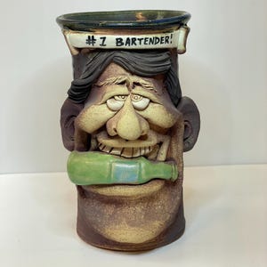 May include: A whimsical ceramic mug shaped like a face, with the text "#1 Bartender!" on a banner. The mug features a green bottle in its mouth and is painted in shades of brown, tan, and black.