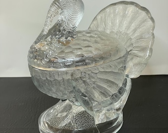 May include: A clear glass turkey-shaped candy dish with a textured surface. The dish features a detailed design of feathers and a curved neck. The base is oval-shaped, and the tail feathers are fanned out. A decorative piece for the home.