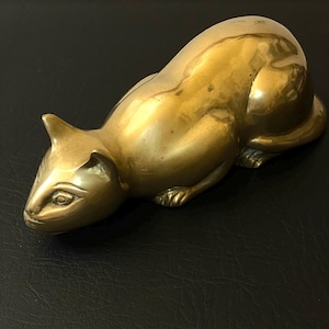 May include: A brass cat figurine in a resting pose. The sculpture has a smooth, polished surface with visible details of the cat's features, including its ears, eyes, and paws. The cat is a warm, golden color.