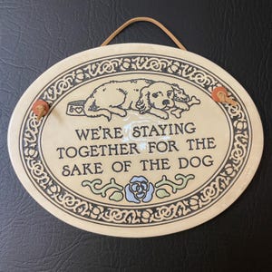 May include: A white ceramic plaque with a black border and a brown rope hanger. The plaque features a cartoon dog and the text "We're staying together for the sake of the dog."