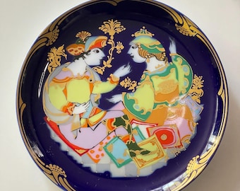 May include: A decorative plate with a dark blue background and a gold rim. The plate features a colourful illustration of two figures in ornate clothing. The figures are surrounded by decorative floral elements.
