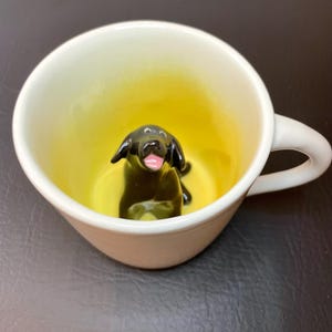 May include: A white ceramic mug with a black dog figurine inside. The interior of the mug is yellow, and the dog has a pink tongue. The mug has a curved handle.