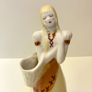 May include: A vintage ceramic figurine of a woman with blonde hair, wearing a white dress with brown accents. The figurine has a basket and closed eyes. The dress features a brown criss-cross design and brown crosses.