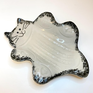 May include: A white ceramic cat-shaped dish with a black speckled rim. The cat's face is drawn with black lines, featuring eyes, a nose, and whiskers. The dish has a wavy design and is suitable for holding small items.