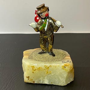 May include: A gold-colored clown figurine with a red and white polka dot bag, a green and gold scarf, and a brown hat. The clown is standing on a white and brown stone base. The base is signed "Romir".