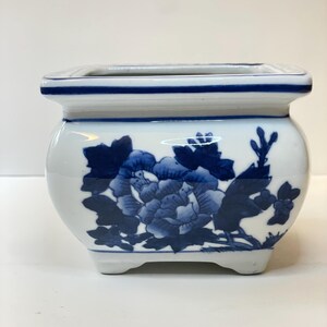 May include: A white ceramic planter with a square top and a rounded body. The planter is decorated with a blue floral design. The rim and base are trimmed with blue. The planter has a small footprint and is suitable for small plants.