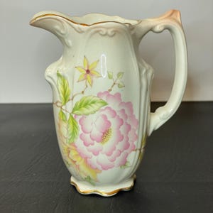 May include: Cream-colored ceramic pitcher with a floral design. The pitcher features a large pink peony, yellow flowers, and green leaves. Gold trim accents the rim and base. The handle is curved and the spout is gently pointed.