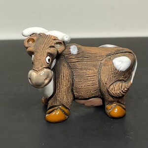 May include: A small, brown ceramic cow figurine with white spots and orange hooves. The cow has a whimsical expression with large eyes and a slightly open mouth.