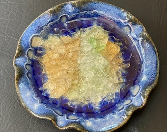 May include: A handmade ceramic ashtray with a blue and brown rim and a deep blue interior. The ashtray contains a mix of colorful, broken glass pieces in shades of yellow, green, and clear.