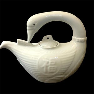May include: White ceramic teapot shaped like a swan with a lid. The teapot has a Chinese character for "fortune" on the front.