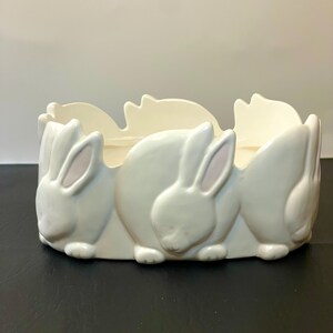 May include: A white ceramic planter with a repeating bunny design. The planter features three-dimensional bunnies around the exterior, with their ears and backs visible. The interior is a smooth, open space for plants or other decorative items.