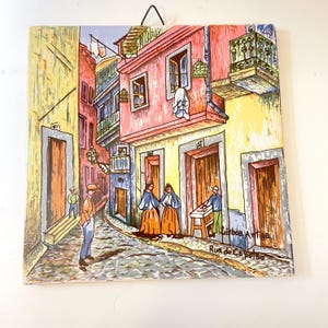 May include: Square ceramic tile featuring a colorful street scene in Lisbon, Portugal. The artwork depicts buildings in shades of pink, yellow, and blue, with figures and cobblestone streets. The tile has a metal wire for hanging.