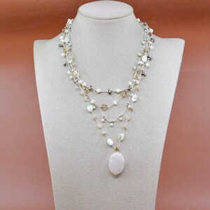 May include: A layered necklace with pearls and beads, presented on a mannequin. The necklace has strands of white, cream, and iridescent beads, with an oval pendant. The background is a plain, muted pink.