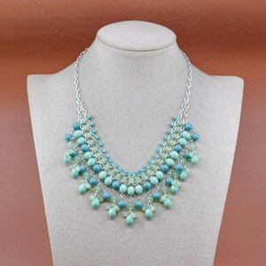 May include: A beaded necklace with a silver chain and multiple strands of turquoise and light green beads. The necklace is displayed on a mannequin bust against a brown background.