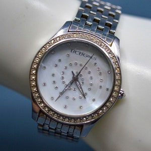 Op de afbeelding: Een zilveren horloge met een witte wijzerplaat en een zilveren band. De wijzerplaat is versierd met kleine strass steentjes en de tekst "BCBGirls" is op de wijzerplaat gedrukt.