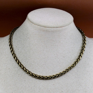 May include: A gold-toned chain necklace with a braided design is displayed on a mannequin. The necklace has a textured appearance and a warm, antique finish. The chain is a choker style, sitting close to the neck.
