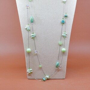May include: A long, delicate necklace with multiple strands, featuring a variety of beads in shades of green, turquoise, and clear. The beads include round, oblong, and stacked shapes, strung on a thin, silver-coloured wire. The necklace is displayed on a neutral-coloured bust.