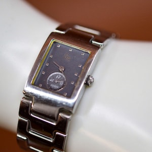 May include: A silver watch with a rectangular face and a black dial. The watch has a silver band and a small sub-dial with the word "Guess" on it.