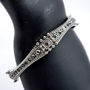 May include: A silver bracelet with a curved design and black gemstones. The bracelet is adorned with intricate details and a square-shaped gemstone in the center.