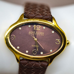 May include: A gold-tone oval watch with a brown leather strap. The watch face is brown with gold-tone hands and Roman numeral hour markers. The watch face also has the text "JOAN RIVERS CLASSICS" and "QUARTZ".