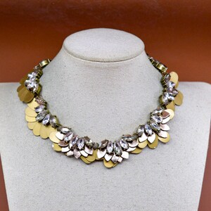 May include: A statement necklace featuring a series of gold-toned metal petal-shaped discs layered with clear, marquise-cut faceted crystals. The design is arranged in a semi-circular collar shape, showcasing a repeating pattern of metallic gold elements.