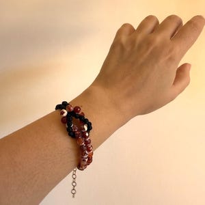Puede incluir: Un brazalete de múltiples hilos con cuentas de ágata rojas y marrones, y cuentas negras. El brazalete se sujeta con una cadena plateada. La joya se lleva en una muñeca sobre un fondo neutro.