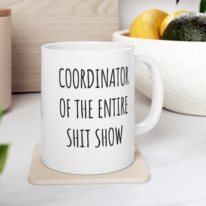 May include: White ceramic coffee mug with black text that reads "Coordinator of the Entire Shit Show". The mug is sitting on a beige coaster.