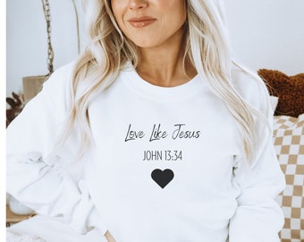 以下が含まれることがあります： 「Love Like Jesus」と「JOHN 13:34」の文字と、その下に黒いハートが描かれた白いスウェットシャツです。
