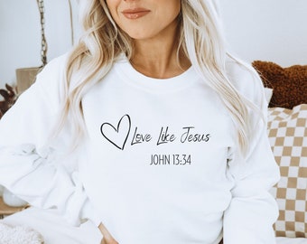 以下が含まれることがあります： 白いスウェットシャツに、黒いハートと「Love Like Jesus」と「JOHN 13:34」の文字がプリントされています。