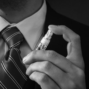 May include: A man in a suit and tie holds a small, clear glass bottle with a white cap. The bottle is partially filled with a liquid.