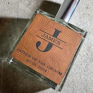 May include: A square glass perfume bottle with a silver cap, featuring a tan leather-like label. The label is engraved with a large letter J, the name JAMES, the text FATHER OF THE GROOM, and the date 07.01.2024 in a dark, bold font.