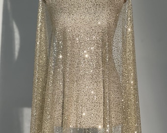 May include: A sheer, beige fabric with a sparkling, glittery pattern is draped over a mannequin. The fabric is semi-transparent, allowing the mannequin's form to be partially visible. The glitter adds a shimmering effect, creating a glamorous look. The fabric is suitable for dresses and other apparel.