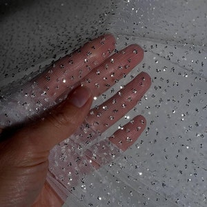 May include: Close-up of white tulle fabric with scattered silver glitter. The sheer material is held up, showing the texture and sparkle of the decorative fabric. Ideal for crafting, fashion, or event decor.