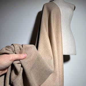 Op de afbeelding: Een beige stof met visgraatpatroon is over een paspop gedrapeerd. De textuur en weefsel van de stof zijn zichtbaar. De stof wordt door een hand vastgehouden. De achtergrond is een effen, lichtgekleurde muur.