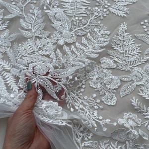 May include: White floral lace fabric with a delicate beaded design. The lace features intricate floral patterns and leaves, creating a romantic and elegant look.