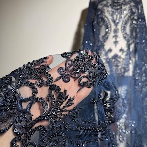 Peut inclure: Gros plan sur un tissu en dentelle bleu marine avec des motifs floraux et tourbillonnants complexes. Le tissu est orné de paillettes et de perles, créant un effet scintillant. Un mannequin drapé dans le même tissu est visible en arrière-plan, mettant en valeur le design.
