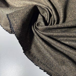 May include: Close-up of a folded fabric with a herringbone pattern. The fabric is a mix of brown and black threads, creating a textured appearance. The edges of the fabric are visible, showing the raw, unfinished cut.