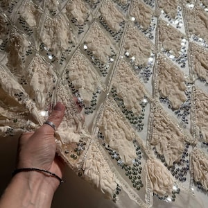 May include: Close-up of a cream fabric with a diamond pattern. Each diamond is decorated with ruffled fabric, silver sequins, and small white beads. The fabric is held by a hand.