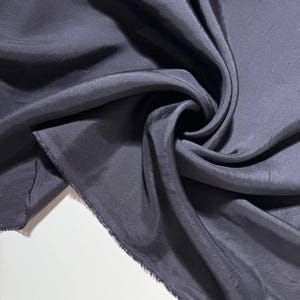 May include: A close-up of a dark gray fabric with a silky sheen. The fabric is draped and folded, creating soft shadows and highlights. The edges of the fabric are unfinished, showing a raw, frayed texture. The material appears to be lightweight.