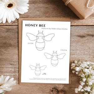 May include: A pattern sheet titled "HONEY BEE" by Nikki Glass Works, featuring three bee designs with dimensions in inches. The sheet is on a wooden surface, with white flowers and a wrapped gift box in the background.