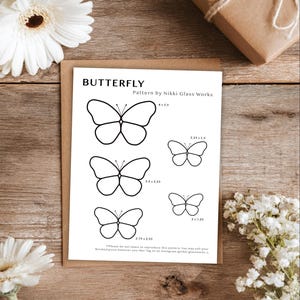 Puede incluir: Una hoja de patrones con diseños de mariposas de varios tamaños, con dimensiones en pulgadas. La hoja se titula "BUTTERFLY" e incluye el texto "Pattern by Nikki Glass Works". La imagen está sobre una superficie de madera, con flores blancas y un regalo envuelto.