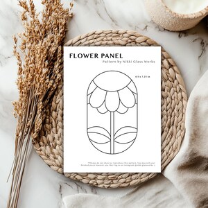 May include: A white paper with a black floral design labeled "FLOWER PANEL" and the text "Pattern by Nikki Glass Works." The dimensions are 4.5 x 7.25 inches. The paper is on a woven placemat with dried wheat stalks.
