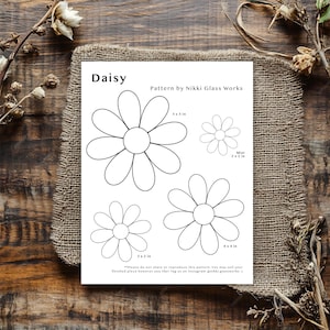May include: A white paper with daisy flower patterns of various sizes, labeled with dimensions in inches. The paper is titled "Daisy" and includes the text "Pattern by Nikki Glass Works". The paper is on a burlap cloth.