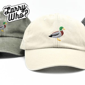 Może przedstawiać: Beżowa czapka z daszkiem z haftowanym kaczką w kolorze zielonym, brązowym i szarym z przodu. Czapka ma zakrzywiony daszek i regulowany pasek z tyłu.