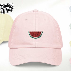 Puede incluir: Una gorra de béisbol rosa con un diseño de sandía bordado. La sandía es roja con semillas negras y una cáscara verde.