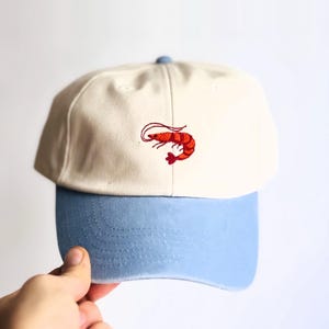 Könnte beinhalten: Eine Baseballkappe mit cremefarbener Krone und hellblauem Schirm. Auf der Vorderseite befindet sich eine kleine, detaillierte Stickerei einer leuchtend orangefarbenen Garnele. Die Kappe hat ein strukturiertes Design mit einem Knopf oben.
