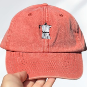 Peut inclure: Casquette de baseball corail avec une image brodée d'une cafetière italienne argentée et noire. La casquette a une visière incurvée et une sangle réglable.